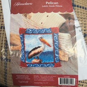 May include: A latch hook pillow kit featuring a pelican design. The kit includes acrylic yarn, a gridded latch hook canvas, and instructions. The finished size is 16 x 16 inches.
