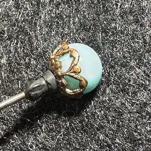 May include: A vintage hat pin with a light blue, round bead. The bead is set in a gold-toned filigree design. The pin has a dark gray, textured base and a silver-colored pin.
