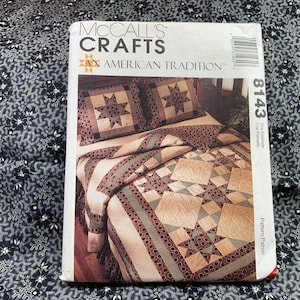 Könnte beinhalten: Ein McCall's Crafts Quilt-Muster mit einem Sternendesign. Das Muster heißt "An American Tradition" und die Musternummer ist 8143. Der Quilt ist aus beigefarbenem Stoff mit roten und grünen Akzenten gefertigt. Der Quilt ist auf einem Bett mit zwei Kissen abgebildet.
