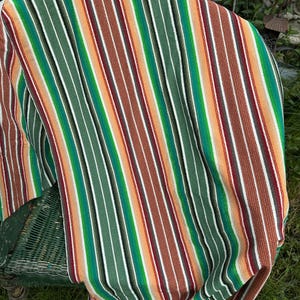 Può includere: Un tessuto a righe colorato, drappeggiato su una sedia di vimini verde. Le righe verticali presentano tonalità di verde, arancione, marrone e bianco. La sedia è parzialmente visibile, aggiungendo consistenza alla composizione.