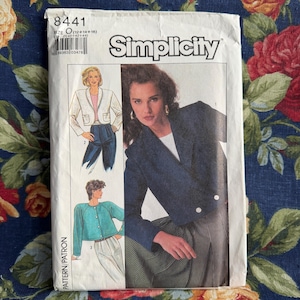 May include: A vintage Simplicity pattern envelope featuring illustrations of women's jackets. The envelope is white with the Simplicity logo and product details. The jackets are in various colors, including white, teal, and navy. The background is a floral fabric.