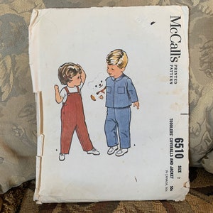 Puede incluir: Patrón de costura vintage McCall's para petos y chaqueta para niños pequeños, talla 2. El patrón presenta un dibujo de línea de dos niños, uno con petos y el otro con una chaqueta. El patrón está impreso en papel blanco con texto azul y negro.