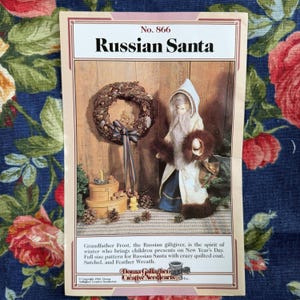 May include: A craft pattern booklet titled No. 866 Russian Santa featuring a photograph of a doll dressed in a white hooded coat with brown fur trim, a blue skirt, and a satchel, alongside a decorative wreath and stacked round boxes.
