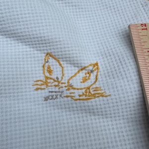 May include: A white fabric kitchen towel with a yellow embroidered design of two birds facing each other.
