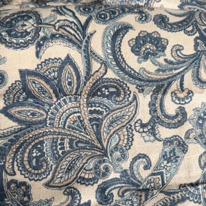 May include: Close-up of a fabric with a paisley pattern in shades of blue, tan, and cream. The design features intricate floral and swirling elements, creating a detailed and ornate textile.