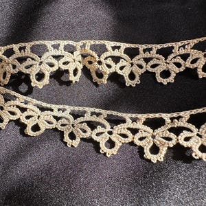 May include: A close-up of two rows of delicate, beige crocheted lace trim. The lace has a repeating pattern of small, intricate loops and knots.