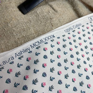 May include: White fabric with a small floral pattern in pink, blue, and green. The fabric has a copyright symbol and the text "Laura Ashley MCMXXXIX" and "100% Cotton" printed on it.