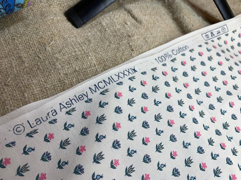 May include: White fabric with a small floral pattern in pink, blue, and green. The fabric has a copyright symbol and the text "Laura Ashley MCMXXXIX" and "100% Cotton" printed on it.