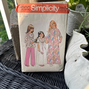 Könnte beinhalten: Vintage Simplicity Schnittmuster Nummer 7202 für Mädchen-Nachthemden und Morgenmäntel. Das Schnittmuster umfasst zwei Größen, 12 und 14. Der Schnittmusterumschlag zeigt Illustrationen von drei Mädchen, die die Kleidungsstücke tragen. Das Nachthemd ist weiß mit rosa Besatz und der Morgenmantel ist rosa mit Patchwork-Muster.