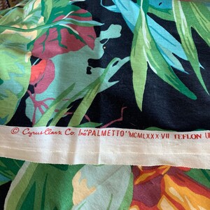 K&ouml;nnte beinhalten: Nahaufnahme eines schwarzen Stoffes mit einem farbenfrohen tropischen Druck. Der Stoff hat ein wei&szlig;es Etikett mit dem Text "Cyrus Clark Co. Inc. 'PALMETTO' MCMILXXXVII TEFLON UP - "