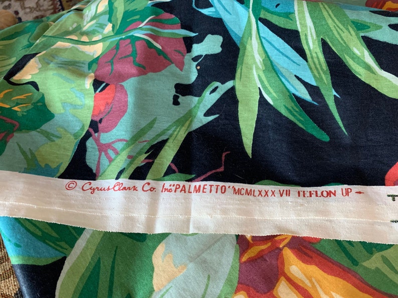 K&ouml;nnte beinhalten: Nahaufnahme eines schwarzen Stoffes mit einem farbenfrohen tropischen Druck. Der Stoff hat ein wei&szlig;es Etikett mit dem Text "Cyrus Clark Co. Inc. 'PALMETTO' MCMILXXXVII TEFLON UP - "