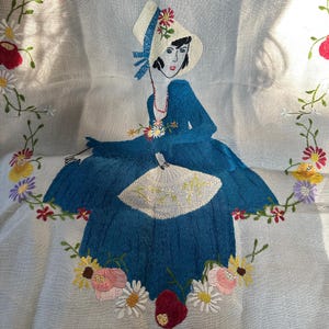May include: A vintage embroidered linen tablecloth featuring a woman in a blue dress and white hat, holding a fan. The tablecloth is decorated with colorful floral embroidery.