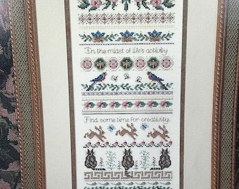 May include: Framed cross-stitch artwork featuring floral and animal motifs. The design includes text that reads "In the midst of life's activity" and "Find some time for creativity." The artwork is framed in a decorative, ornate frame.