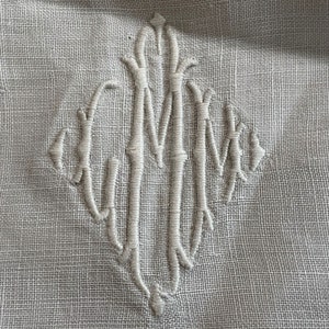 May include: A white embroidered monogram on a linen fabric background. The monogram is a stylized "MM" with the letters intertwined.