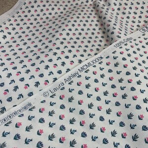 May include: White fabric with a repeating pattern of small pink flowers, green leaves, and blue berries. The fabric has a label that reads "Laura Ashley MCMILXXXIX 100% Cotton".