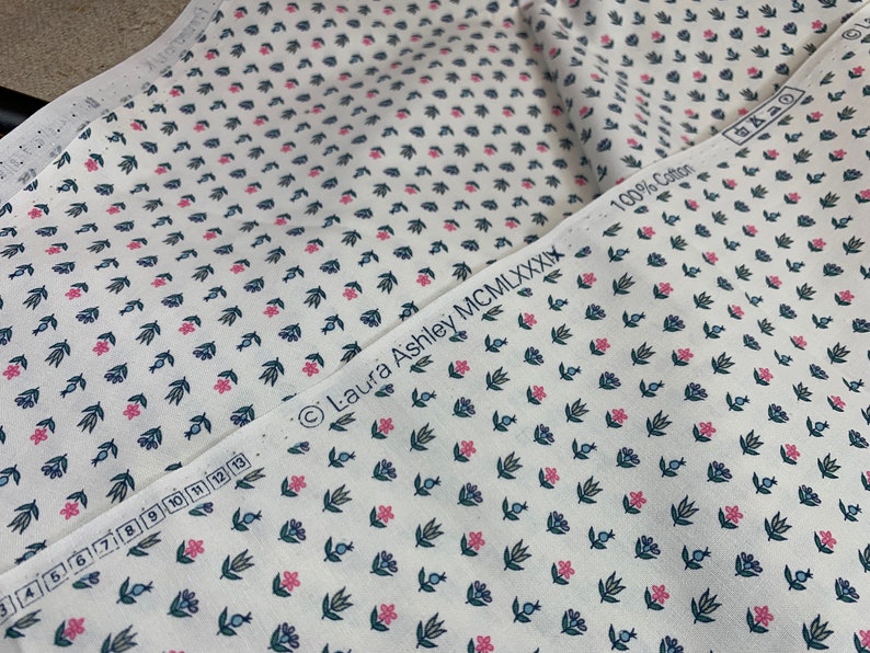 May include: White fabric with a repeating pattern of small pink flowers, green leaves, and blue berries. The fabric has a label that reads "Laura Ashley MCMILXXXIX 100% Cotton".