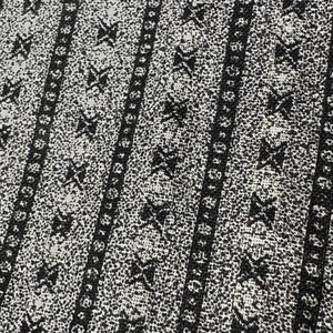 May include: Close-up of a fabric with a repeating pattern. The design features black, stylized cross-like shapes and vertical stripes on a speckled white background. The stripes are bordered by small, circular shapes.