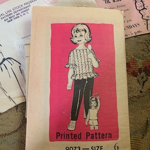 Könnte beinhalten: Ein Vintage-Schnittmuster für ein Mädchenkleid. Das Schnittmuster ist auf rosa Hintergrund gedruckt und zeigt eine Schwarzweißillustration eines Mädchens, das ein Kleid mit Rüschenkragen und schwarze Hose trägt. Das Schnittmuster ist mit "Gedrucktes Muster 9073 - Größe 6" beschriftet.