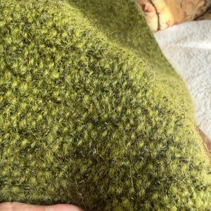 May include: Close-up of a textured, olive green fabric. The material has a dense, looped pile, with a slightly uneven surface. The fabric appears to be a wool blend, with a soft, fuzzy texture. The color is a muted, earthy green.