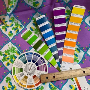 Pu&ograve; includere: Una ruota dei colori, un righello e diversi campioni di colore Pantone sono disposti su un tessuto viola e bianco con un motivo a trifoglio. I campioni sono in tonalit&agrave; di blu, verde, viola e arancione.