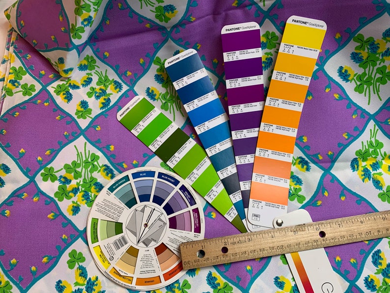 Pu&ograve; includere: Una ruota dei colori, un righello e diversi campioni di colore Pantone sono disposti su un tessuto viola e bianco con un motivo a trifoglio. I campioni sono in tonalit&agrave; di blu, verde, viola e arancione.