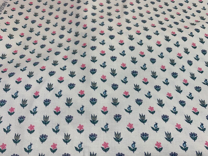 May include: White fabric with a repeating pattern of small pink and blue flowers with green stems. The fabric has a subtle texture.
