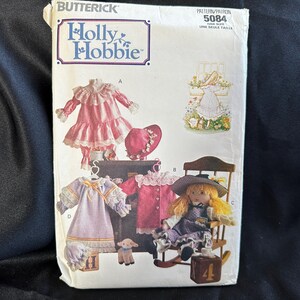 May include: A vintage sewing pattern for a doll dress, featuring a pink dress with lace trim, a purple dress with lace trim, and a doll in a purple dress sitting in a rocking chair. The pattern is from Butterick, Holly Hobbie, and is pattern number 5084.