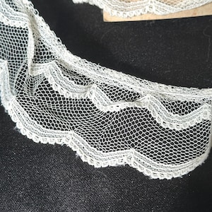 May include: Close-up of delicate white lace trim with a scalloped edge. The lace features a fine, open net pattern, creating a textured and airy appearance. The trim is likely used for embellishing garments or craft projects.