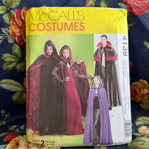 Pode incluir: Um envelope de molde de costura McCall's Costumes, número 4139, com ilustrações de três capas e mantos com capuz diferentes em preto, vermelho e roxo. A embalagem amarela e verde exibe as variações de fantasia A, B e C.