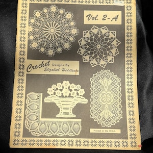 Op de afbeelding: Een vintage instructieboekje getiteld Vol. 2-A Crochet Designs By Elizabeth Hiddleson. De omslag heeft een donkergrijze achtergrond met crèmekleurige illustraties van diverse ingewikkelde gehaakte kleedjes en een bloemenmandpatroon.