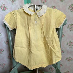 May include: A yellow baby dress with a white lace collar and embroidered details. The dress has short sleeves and a gathered skirt.
