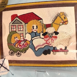 May include: Embroidered artwork featuring a doll with yellow hair, a baby in a green carriage, a house with a red roof, a yellow horse, and a sheep with a pink bow. The artwork is framed in a brown wooden frame.