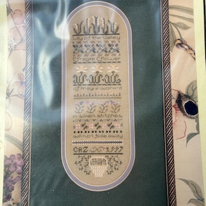 Pode incluir: Um sampler de ponto cruz emoldurado com motivos florais e o texto "Lily of the Valley" e "Fragile Flower". O sampler inclui o texto "of may a work'd" e "in silken stitches will not fade away". A data de 1997 é visível.