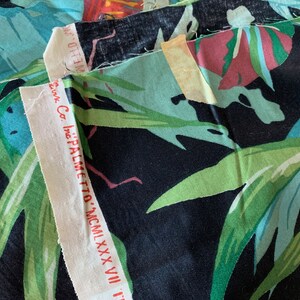 K&ouml;nnte beinhalten: Nahaufnahme eines Stoffmusters mit einem tropischen Blattdruck in Gr&uuml;n-, Blau- und Rosat&ouml;nen auf schwarzem Hintergrund. Der Stoff ist mit "PALMETTO - MCMXXXVII" und dem Text "Inc. Co." beschriftet.
