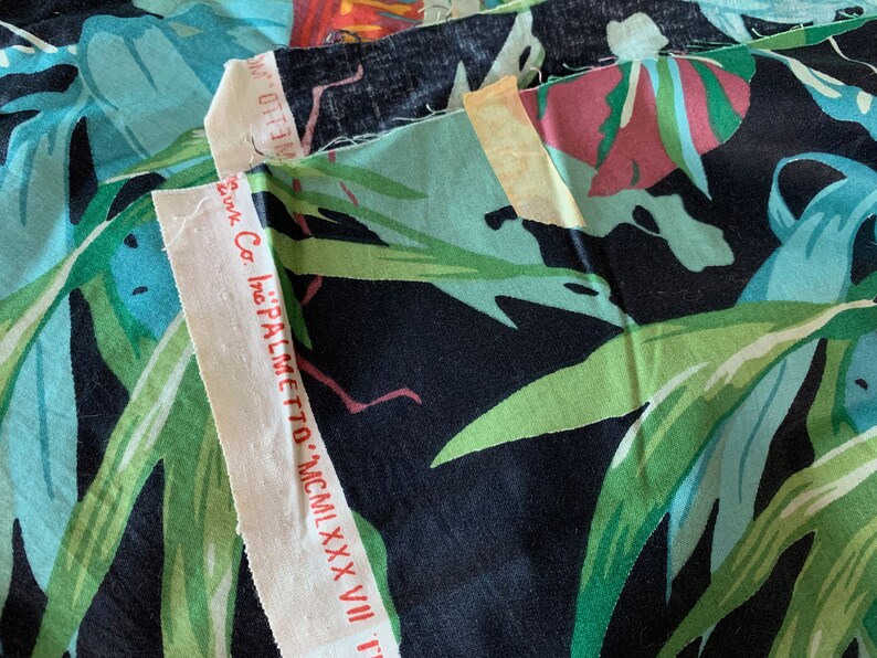 K&ouml;nnte beinhalten: Nahaufnahme eines Stoffmusters mit einem tropischen Blattdruck in Gr&uuml;n-, Blau- und Rosat&ouml;nen auf schwarzem Hintergrund. Der Stoff ist mit "PALMETTO - MCMXXXVII" und dem Text "Inc. Co." beschriftet.