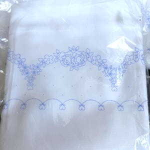 Può includere: Tessuto bianco con ricamo floreale blu. Il ricamo presenta un motivo a ghirlanda floreale nella parte superiore e un bordo smerlato nella parte inferiore. Il tessuto è confezionato in plastica trasparente.