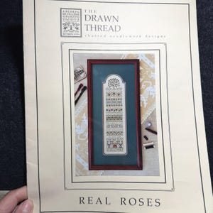 Peut inclure: Un modèle de broderie encadré intitulé "Real Roses" par The Drawn Thread. Le motif présente un motif vertical avec des motifs floraux et géométriques dans des tons de rouge, vert et marron. Le cadre est marron foncé et le fond est vert.