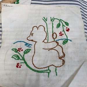 Pode incluir: Um quadrado de tecido branco com bordado marrom, verde e vermelho de um urso sentado em um galho com folhas e bagas. O tecido tem a palavra "three" escrita nele.