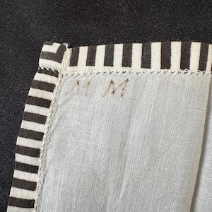 May include: A white fabric with black stripes along the edge. The fabric has the letters "NM" written on it in brown ink.