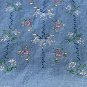 Könnte beinhalten: Ein hellblauer Stoff mit gestickten weißen, rosa und grünen Blumen. Der Stoff hat einen zarten, Vintage-Look.