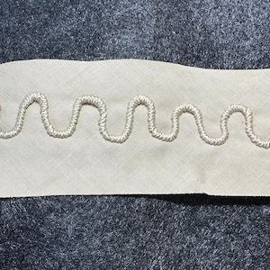 May include: A close-up of a white fabric trim with a wavy, stitched design. The trim is on a dark gray background.