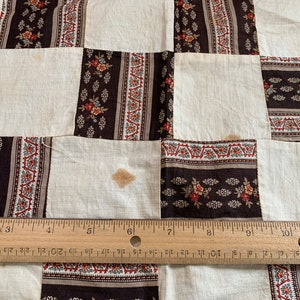 May include: A white quilt with a brown and white floral pattern. The quilt is made of squares and is sewn together with a brown thread. The quilt is measured with a ruler.