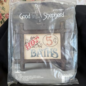 May include: A cross-stitch kit by Good Shepherd, featuring a framed design that reads "HOT BATHS" with a price of 5¢. The kit includes stamped fabric, needle, directions, and floss. The approximate size is 5" x 7".