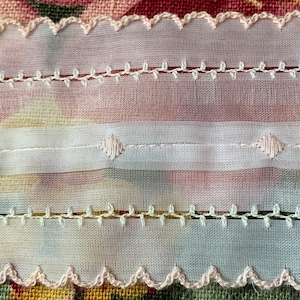 May include: A close-up of white lace trim with pink scalloped edges. The lace has a delicate, embroidered design with small, evenly spaced stitches.