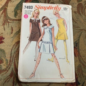 May include: Vintage Simplicity sewing pattern number 7493 for a young junior or teen girl's dress. The pattern includes three different styles: a brown dress with a white collar, a yellow dress with a white collar, and a blue dress with a white collar and white polka dots. The pattern is for sizes 9-10 and has a bust measurement of 77.5 cm.