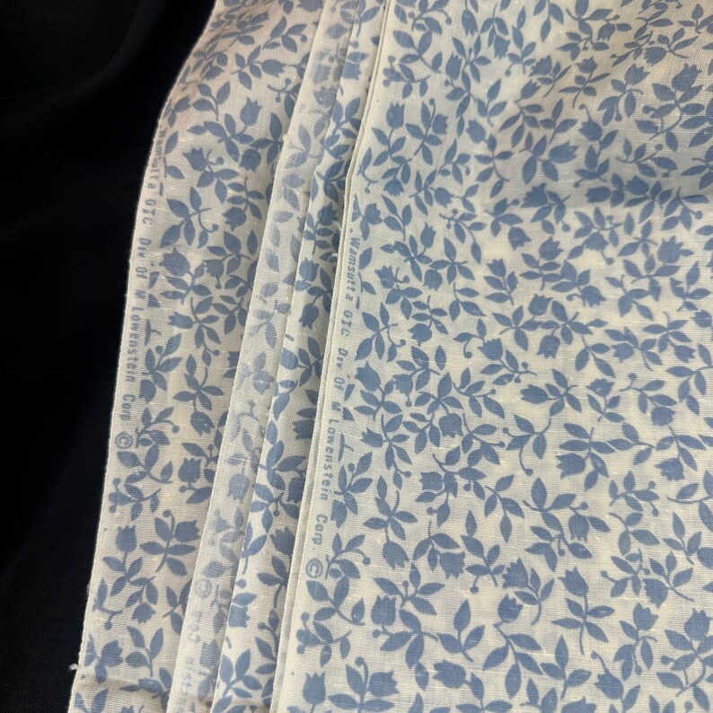 May include: A stack of white fabric with a blue floral pattern. The fabric has the text "Wm.sutta Div. of M. Lowenstein Corp." printed on the side.
