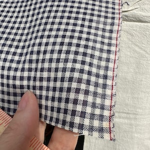Op de afbeelding: Een close-up van een witte en blauwe gingham stof met een rode gestikte rand. De stof is geweven met een fijn geruit patroon.