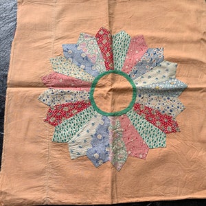 May include: A vintage quilt square with a peach background and a large, multi-colored flower design. The flower is made of fabric scraps in shades of pink, blue, green, and white, with a green circle in the center.