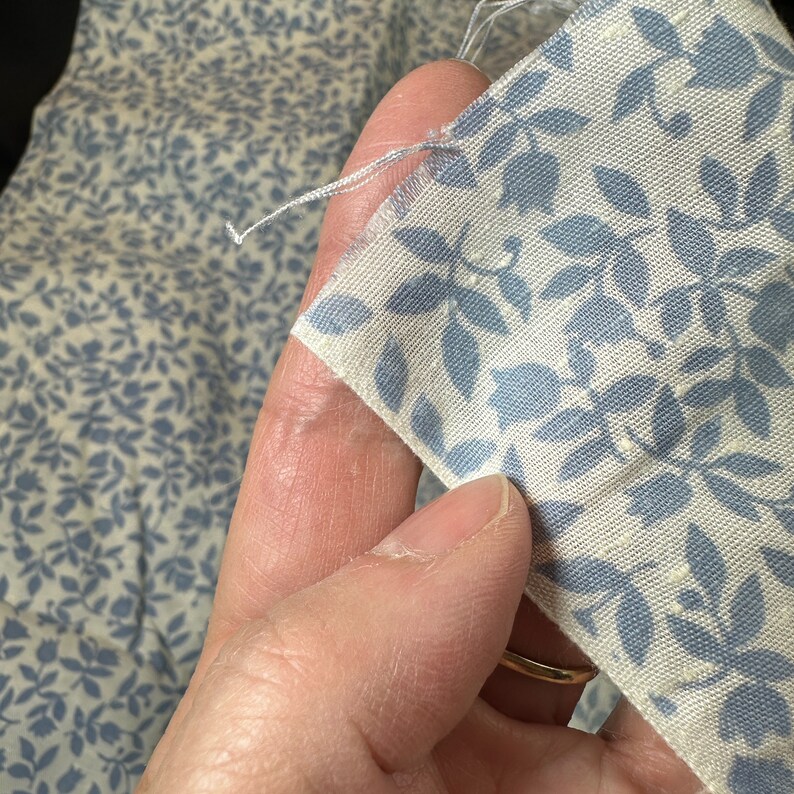 May include: A close-up of a white fabric with a blue floral pattern. The fabric is being held by a hand.