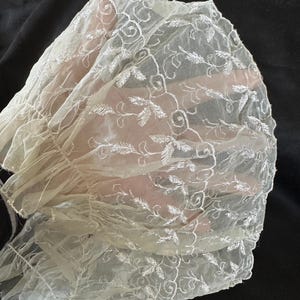 May include: A white sheer fabric with delicate embroidered floral designs. The fabric is gathered and ruffled, creating a soft and romantic look.